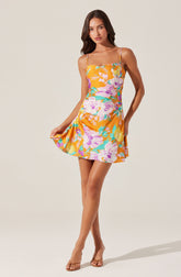 A young woman is wearing a sleeveless, floral print dress with thin spaghetti straps. The dress features a vibrant pattern of large pink, purple, yellow, and green flowers on an orange background. She is also wearing open-toe, nude-colored high-heeled sandals.